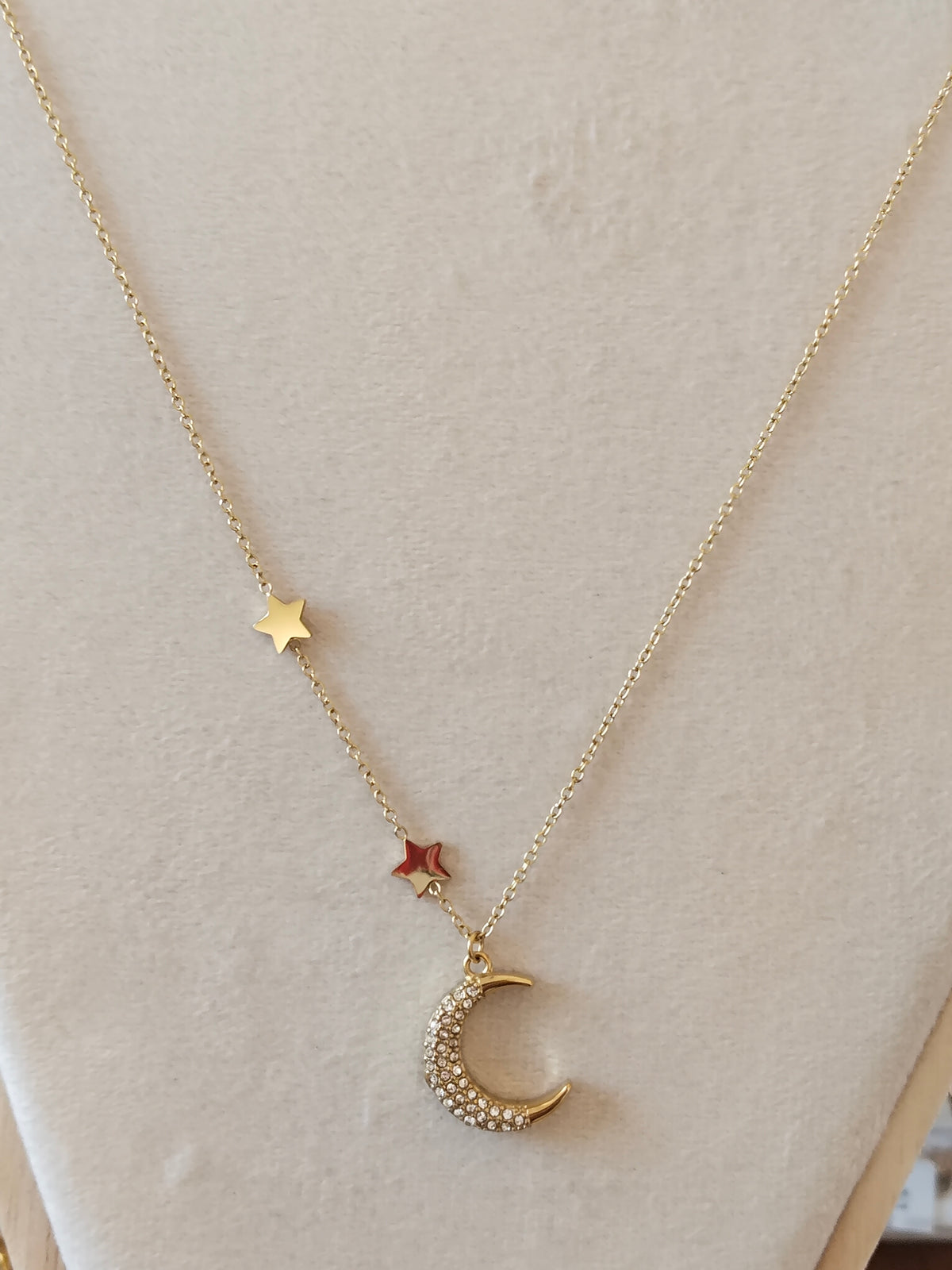 A delicate gold necklace displayed on a cream-colored jewelry bust. The necklace features a fine chain with two gold stars, one small red star, and a crescent moon charm embellished with tiny clear rhinestones.