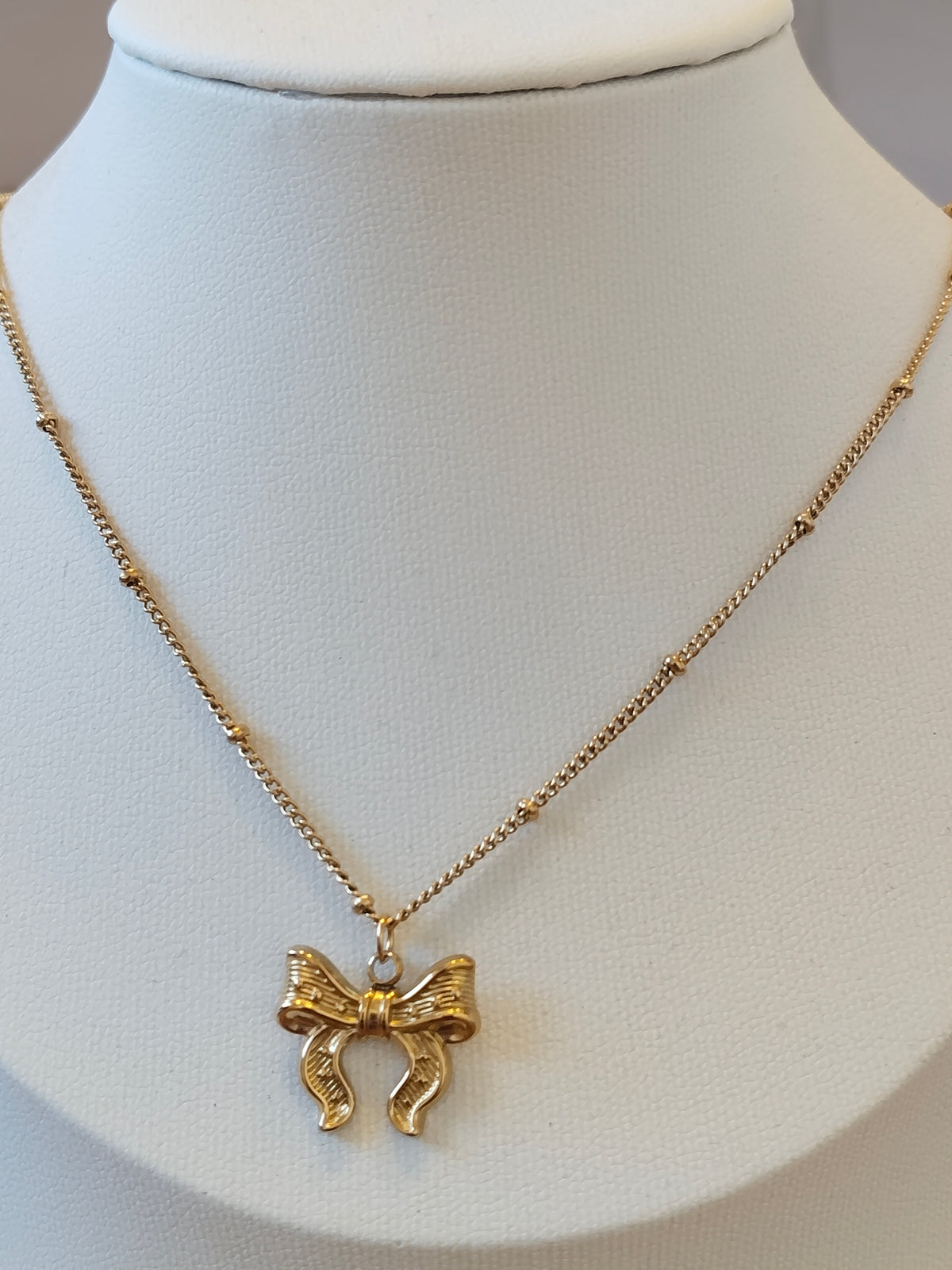 A gold necklace with a textured bow pendant displayed on a white neck mannequin.