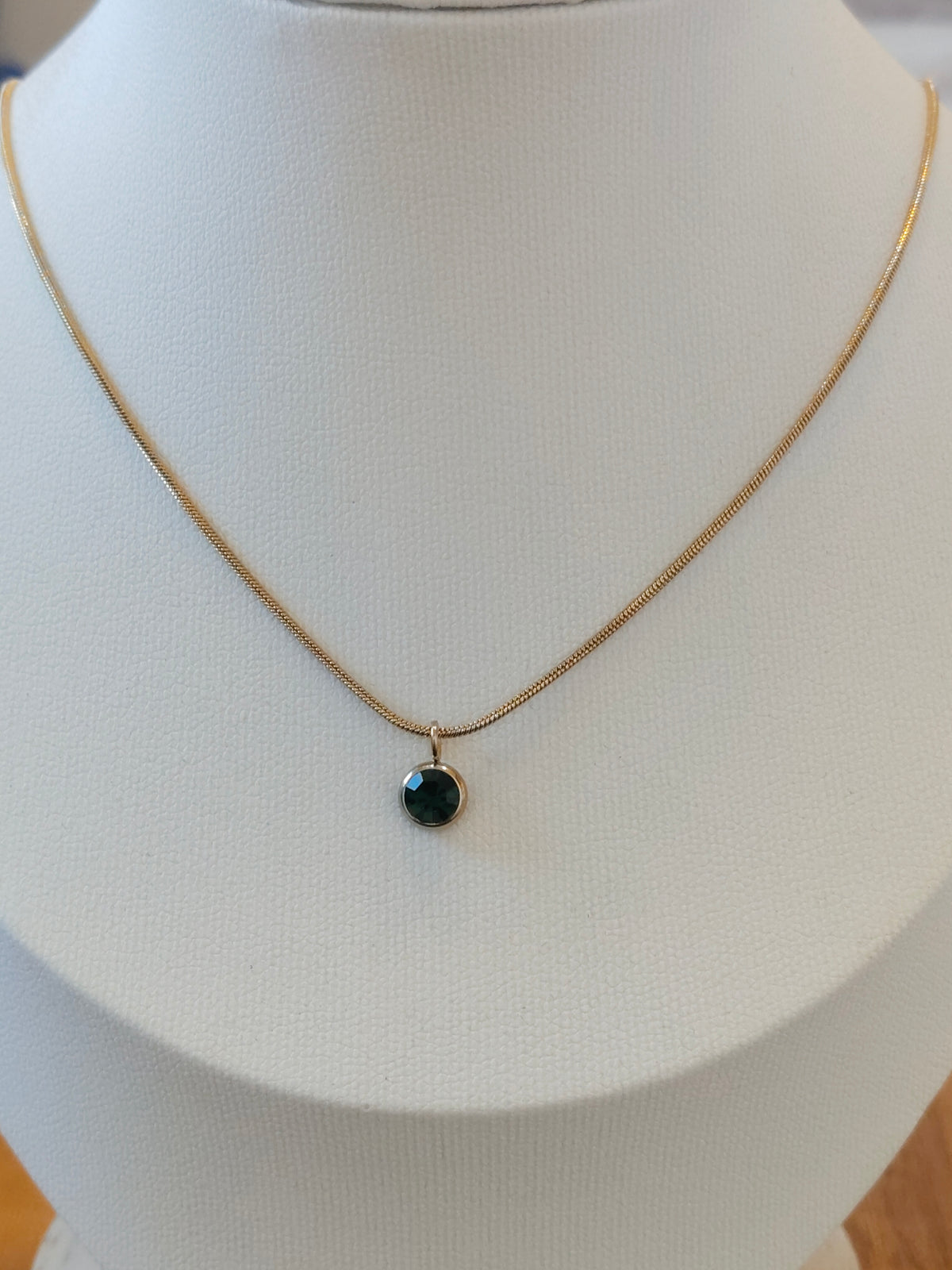 A delicate gold chain necklace with a small round dark green gemstone pendant, displayed on a white jewelry bust.