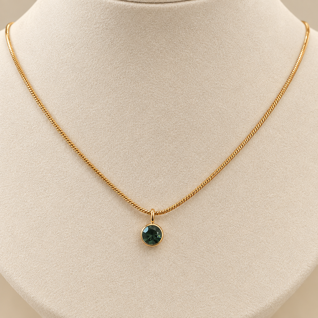 A delicate gold chain necklace with a small round dark green gemstone pendant, displayed on a cream-colored jewelry bust.