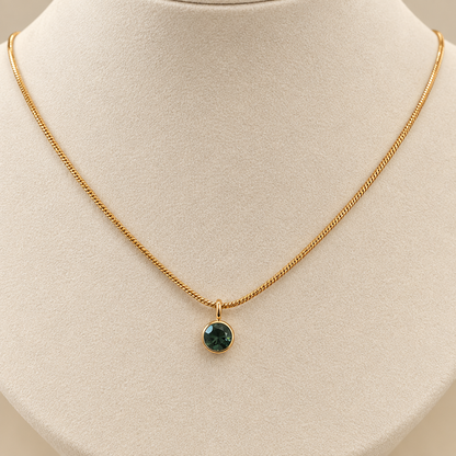 A delicate gold chain necklace with a small round dark green gemstone pendant, displayed on a cream-colored jewelry bust.