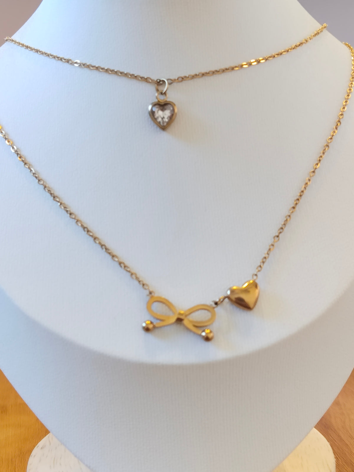 A gold-toned layered necklace displayed on a white jewelry bust. The necklace features a delicate chain with a small heart-shaped pendant and a longer chain with an infinity symbol and a heart charm.