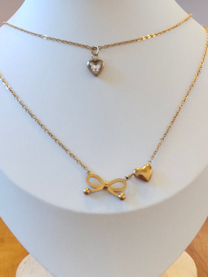 A gold-toned layered necklace displayed on a white jewelry bust. The necklace features a delicate chain with a small heart-shaped pendant and a longer chain with an infinity symbol and a heart charm.