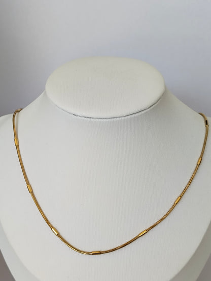 A gold snake chain necklace with small cylindrical links, displayed on a white fabric jewelry bust against a light gray background.