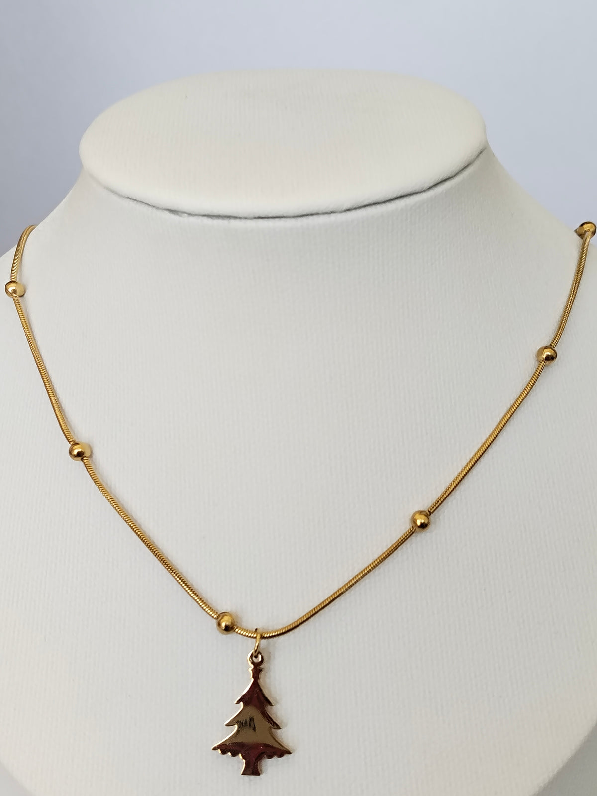 A gold chain necklace with small bead accents and a small Christmas tree pendant, displayed on a white neck mannequin.