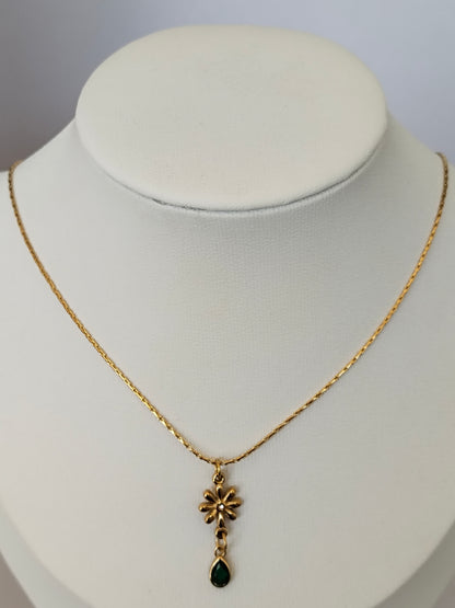A gold chain necklace with a small flower-shaped pendant and a dangling dark green teardrop stone, displayed on a white jewelry bust.