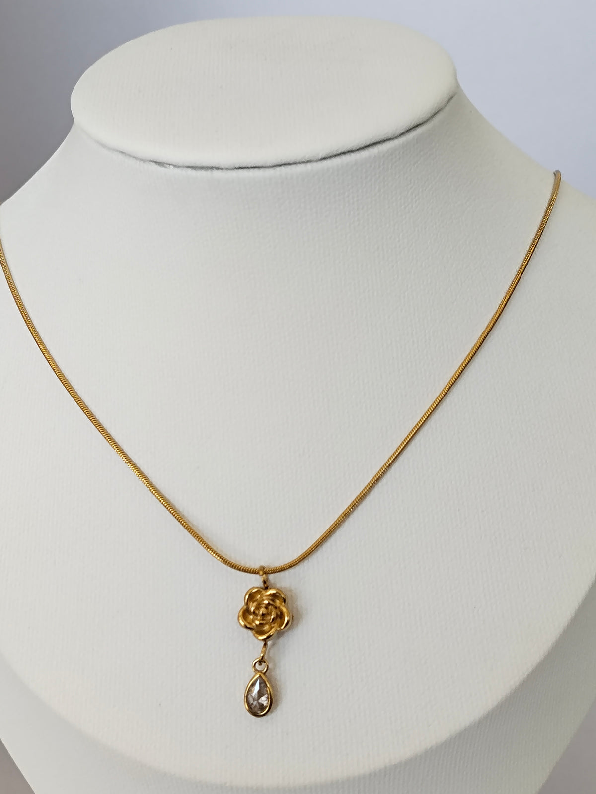 A gold chain necklace with a rose-shaped pendant and a dangling teardrop gemstone, displayed on a white jewelry bust.