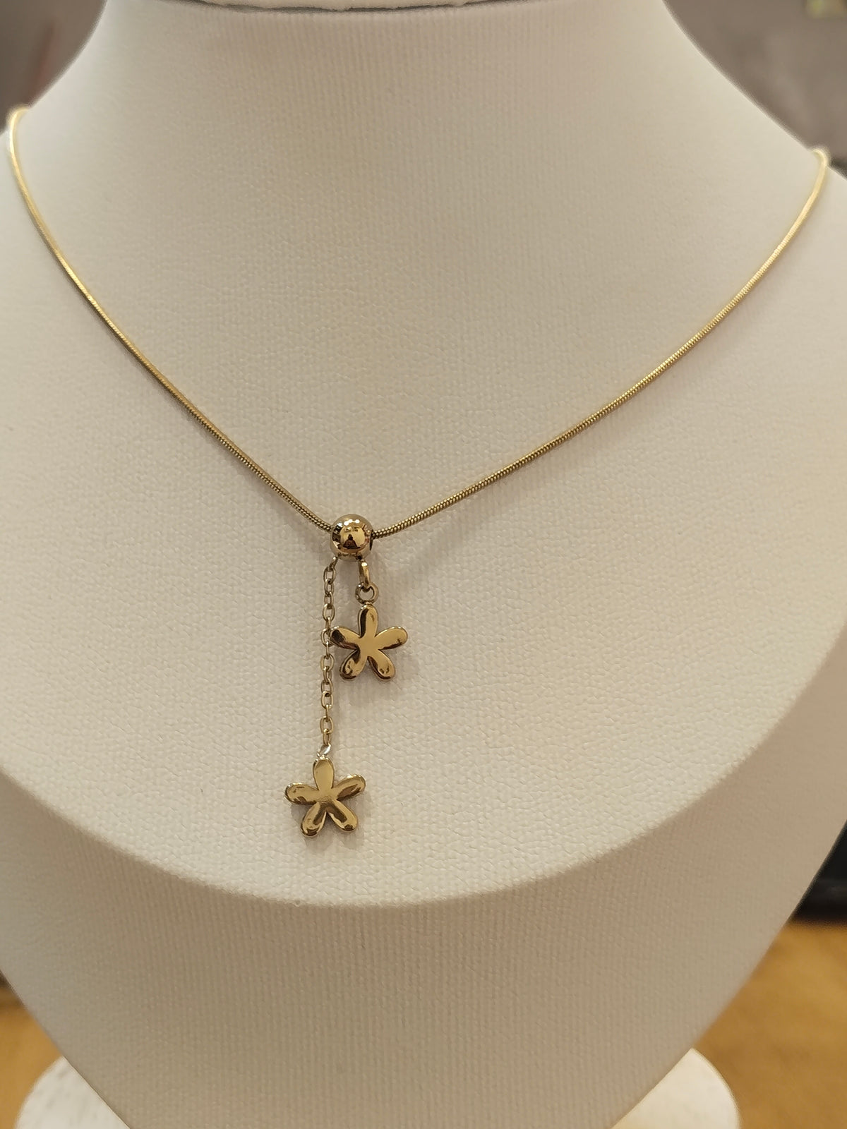 A gold chain necklace displayed on a cream-colored jewelry bust, featuring a central charm with two dangling flower-like charms.