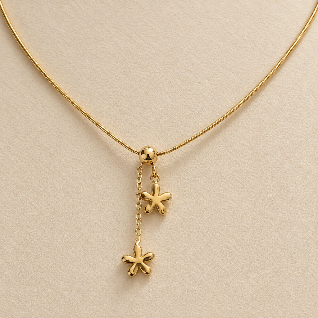 A gold chain necklace displayed on a cream-colored jewelry bust, featuring a central charm with two dangling flower-like charms.