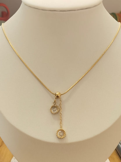 A delicate gold chain necklace displayed on a cream-colored jewelry bust. The necklace features a small gold clasp with two dangling circular charms, each adorned with a clear stone surrounded by a gold textured border.