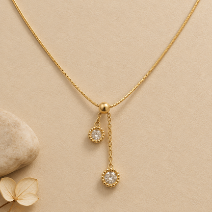 A delicate gold chain necklace displayed on a cream-colored jewelry bust. The necklace features a small gold clasp with two dangling circular charms, each adorned with a clear stone surrounded by a gold textured border.