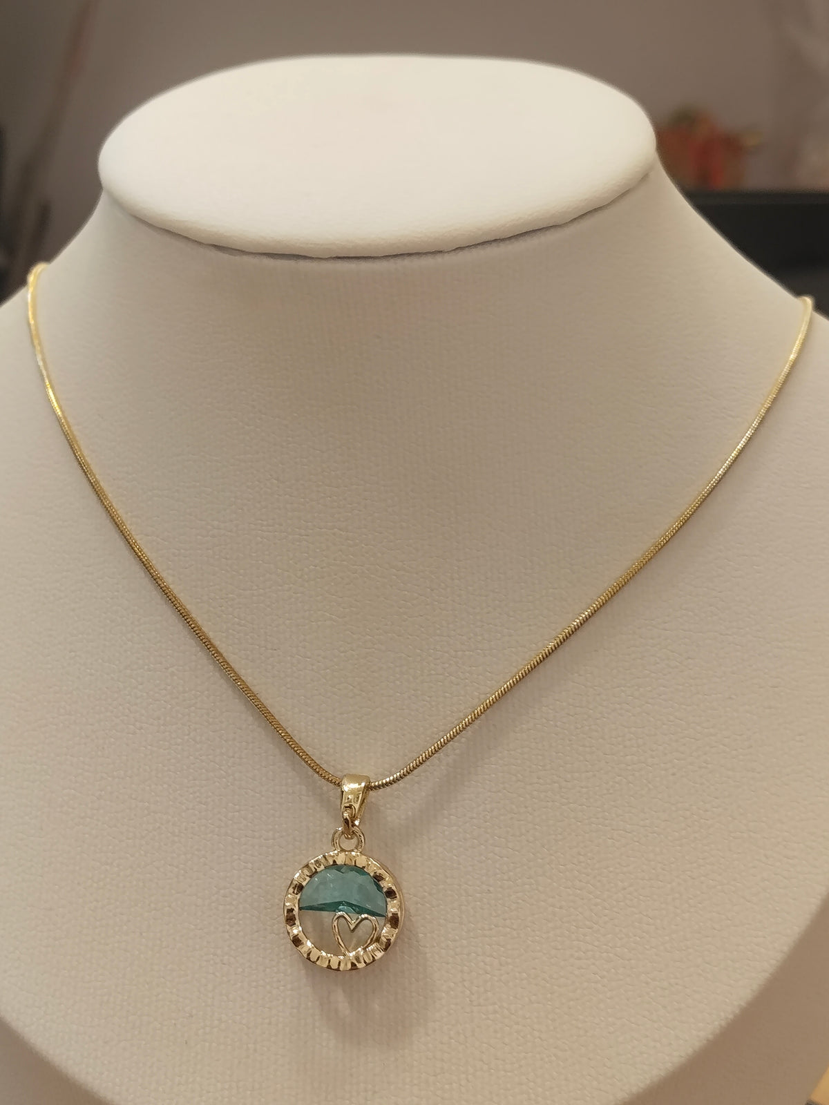 A gold chain necklace with a circular pendant displayed on a cream-colored jewelry bust. The pendant features a turquoise blue and white mountain design surrounded by a border of small clear stones.