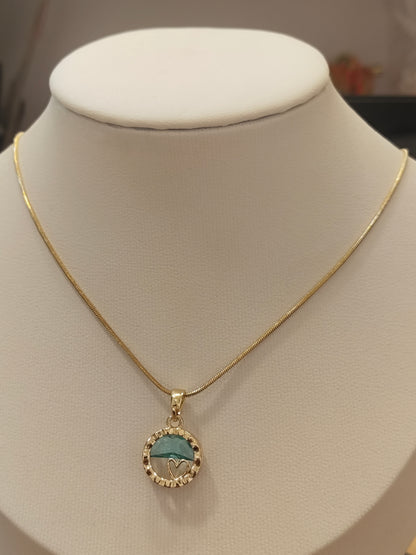 A gold chain necklace with a circular pendant displayed on a cream-colored jewelry bust. The pendant features a turquoise blue and white mountain design surrounded by a border of small clear stones.