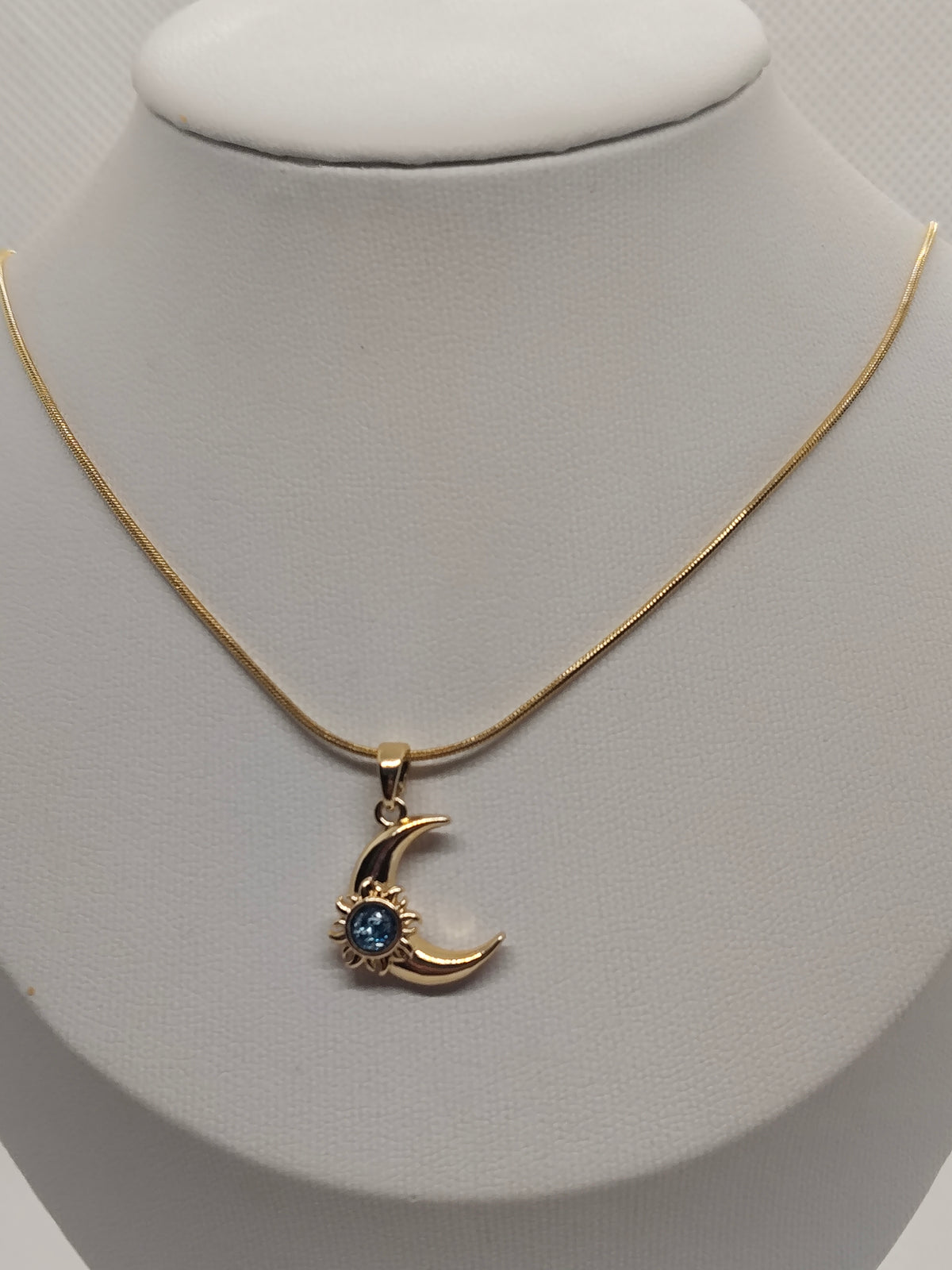 A gold chain necklace with a crescent moon pendant featuring a small blue gemstone, displayed on a white jewelry bust.
