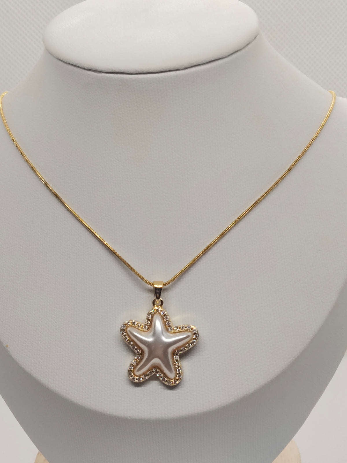 A gold chain necklace with a starfish-shaped pendant, displayed on a white jewelry bust. The pendant features a pearlescent white center surrounded by a gold border with small clear stones.