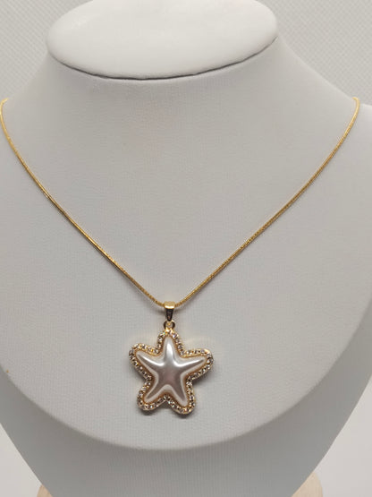 A gold chain necklace with a starfish-shaped pendant, displayed on a white jewelry bust. The pendant features a pearlescent white center surrounded by a gold border with small clear stones.