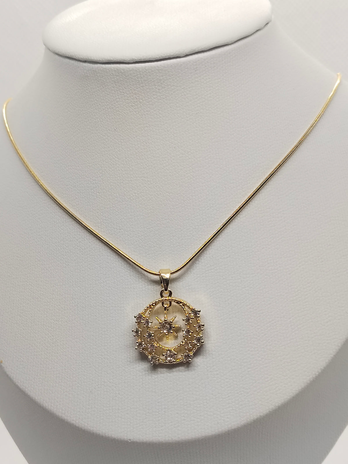 A gold chain necklace with a circular pendant featuring a central sunburst design and surrounding clear stones, displayed on a white neck mannequin.