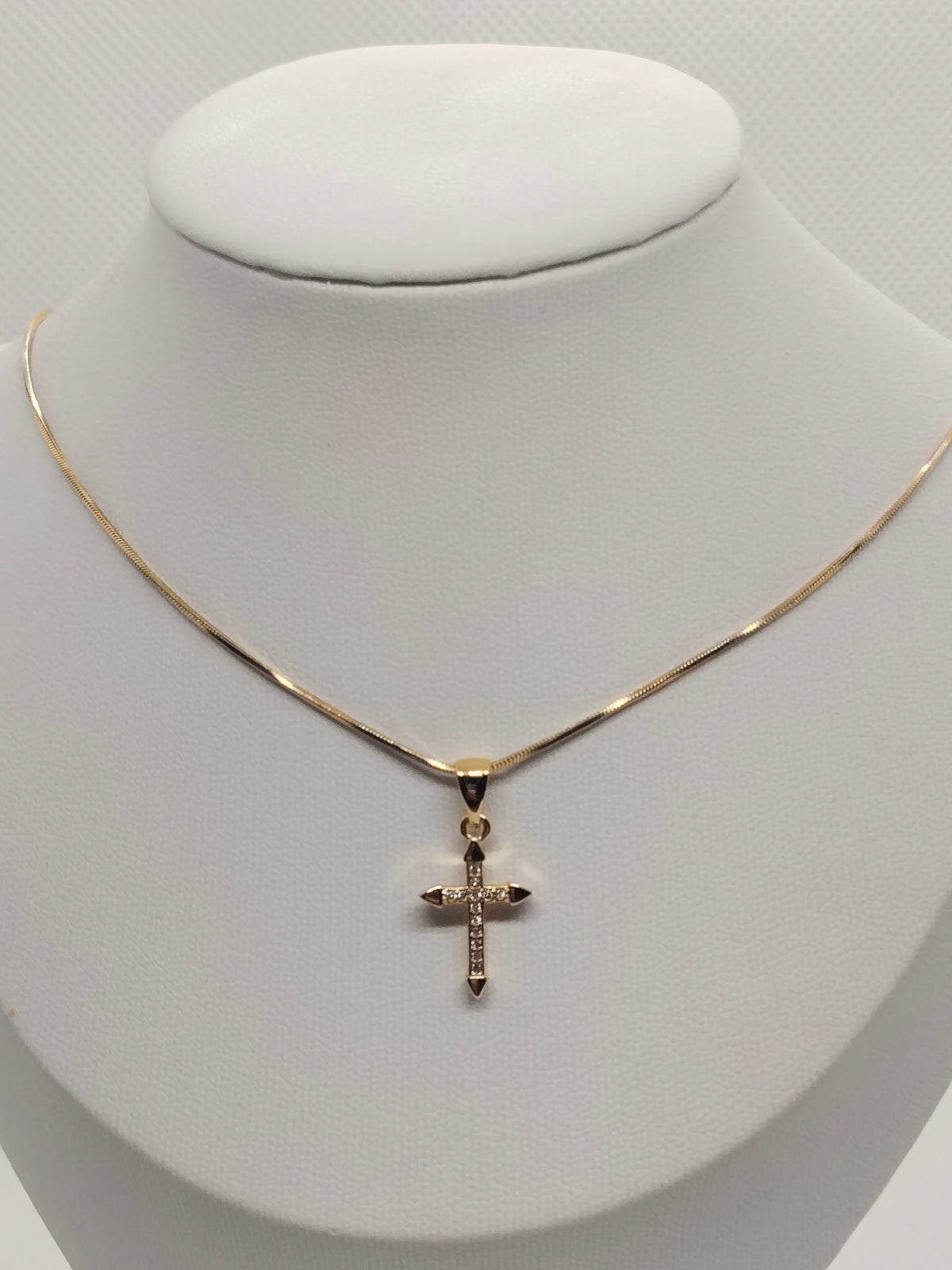 A delicate gold chain necklace with a small cross pendant, displayed on a white jewelry bust.