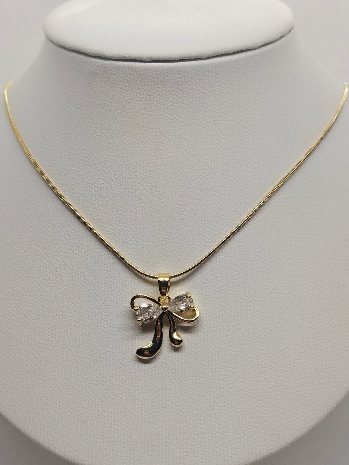 A gold necklace with a bow-shaped pendant set with clear rhinestones, displayed on a white jewelry bust.