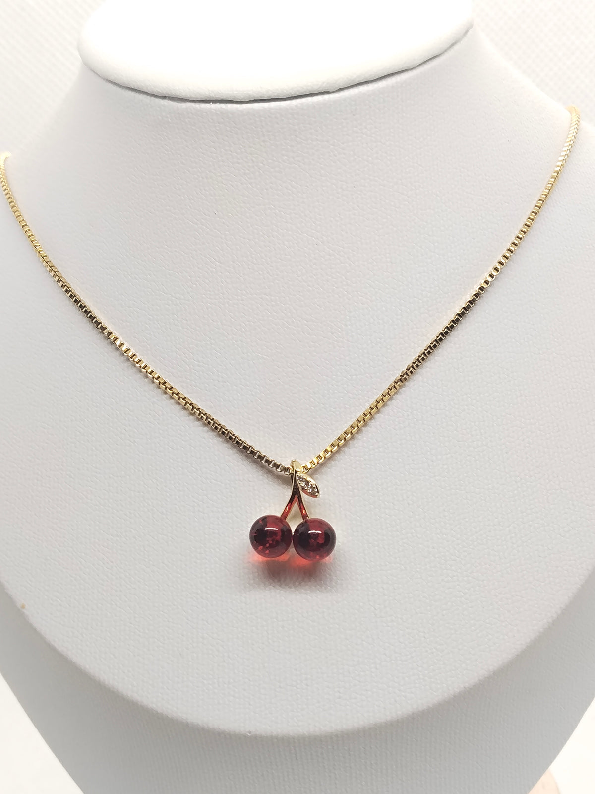 A gold chain necklace with a double cherry pendant made of dark red beads, displayed on a white jewelry bust.