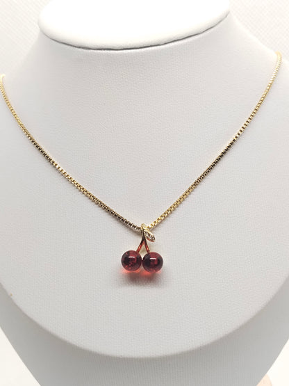 A gold chain necklace with a double cherry pendant made of dark red beads, displayed on a white jewelry bust.