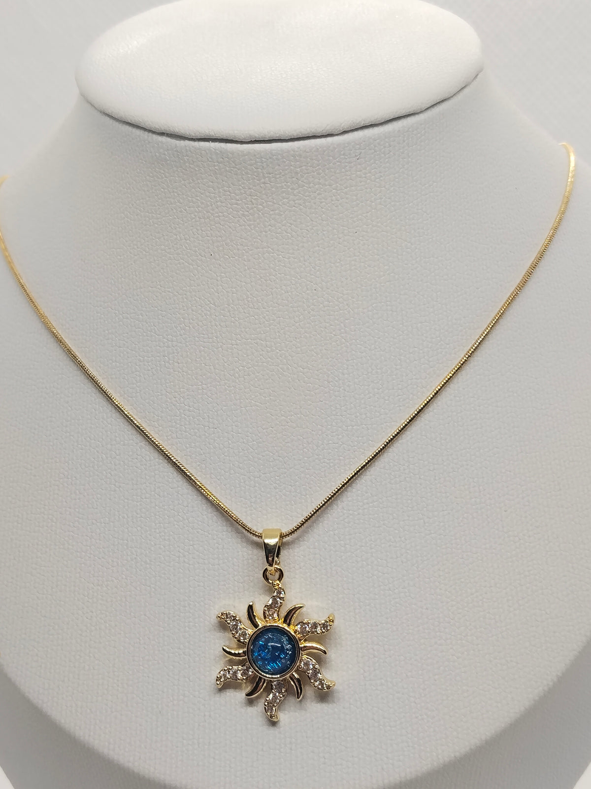 A gold chain necklace with a sunburst pendant featuring a central blue gemstone and small clear stones, displayed on a white jewelry bust.