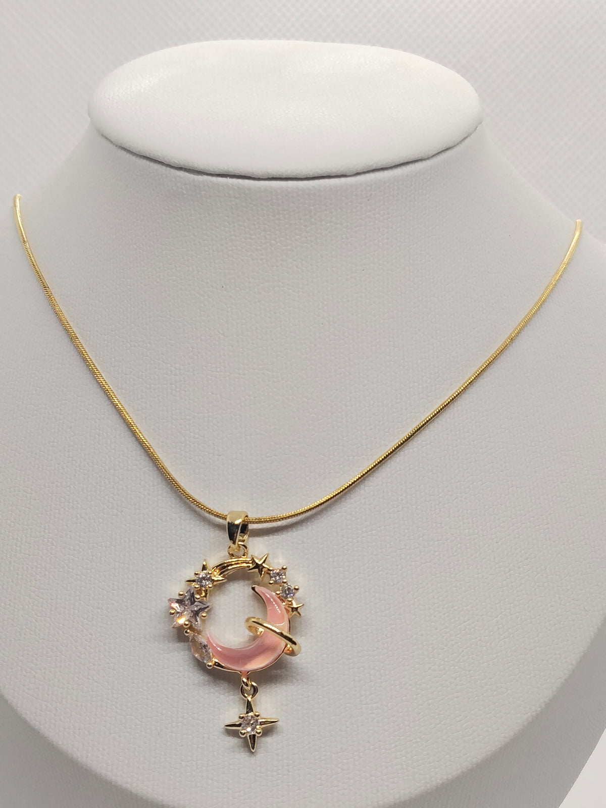 A gold chain necklace with a celestial pendant featuring a pink crescent moon, small clear crystals, and a dangling gold star, displayed on a white mannequin bust.
