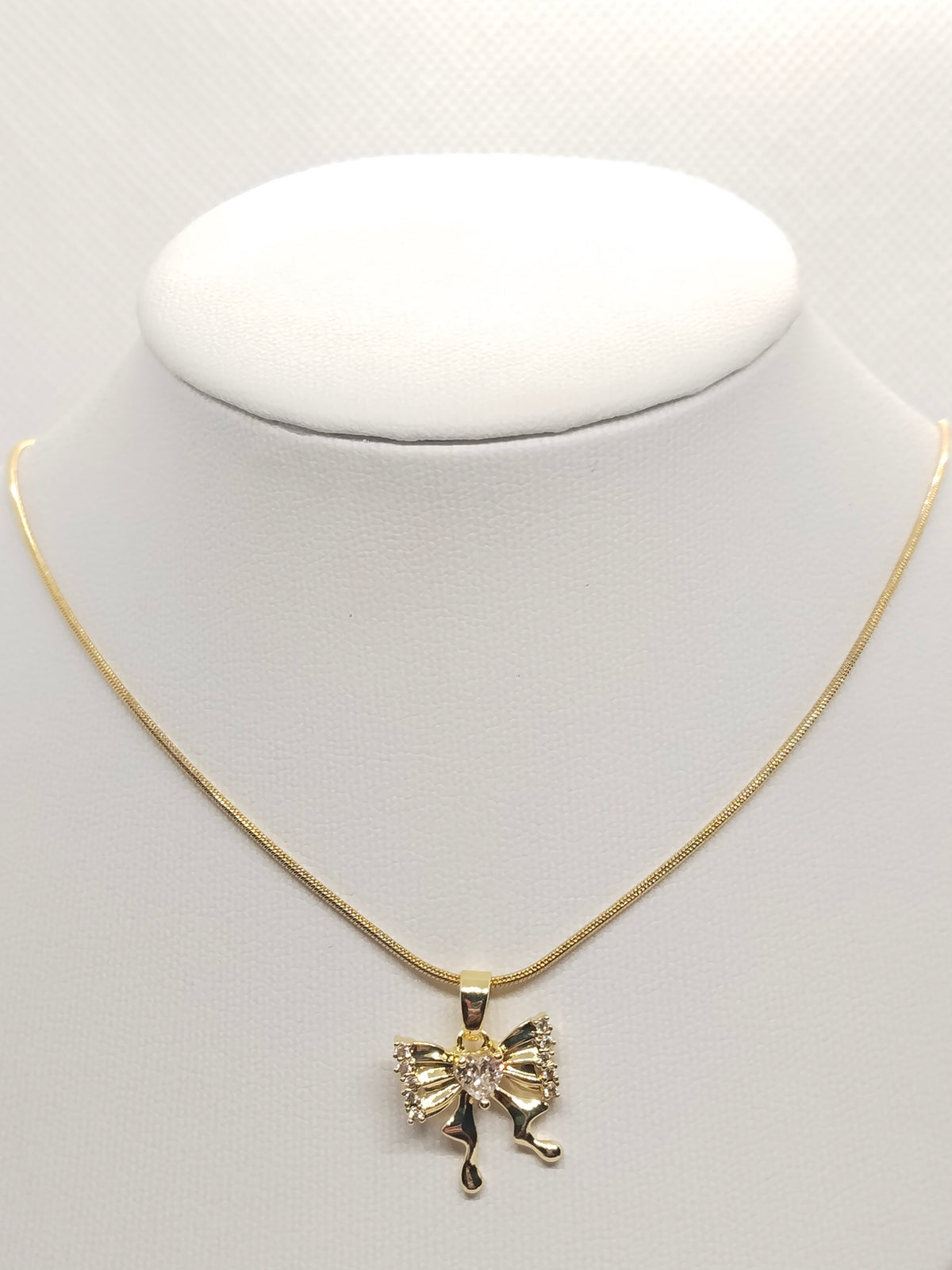 A gold chain necklace with a bow-shaped pendant featuring a central clear rhinestone, displayed on a white jewelry bust against a light gray background.