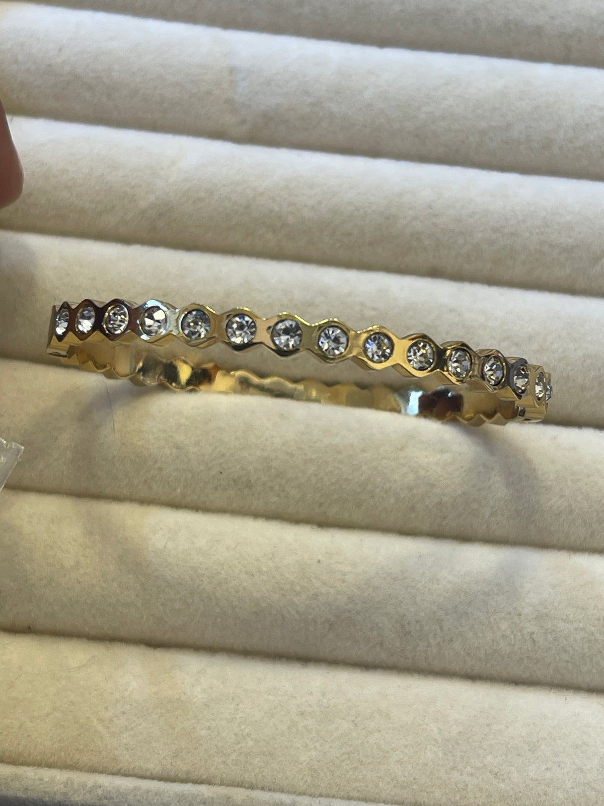 A gold bangle bracelet with a row of clear round-cut gemstones, displayed on a cream-colored jewelry cushion.