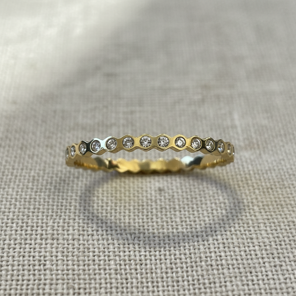 A gold eternity ring featuring a row of small clear round-cut stones set in a honeycomb pattern, resting on a textured beige linen fabric.