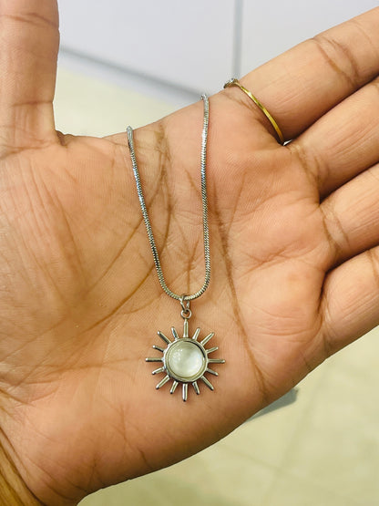 A silver sun pendant necklace with a round white stone, displayed on an open hand. The necklace is shown alongside a gold ring on the model's finger.