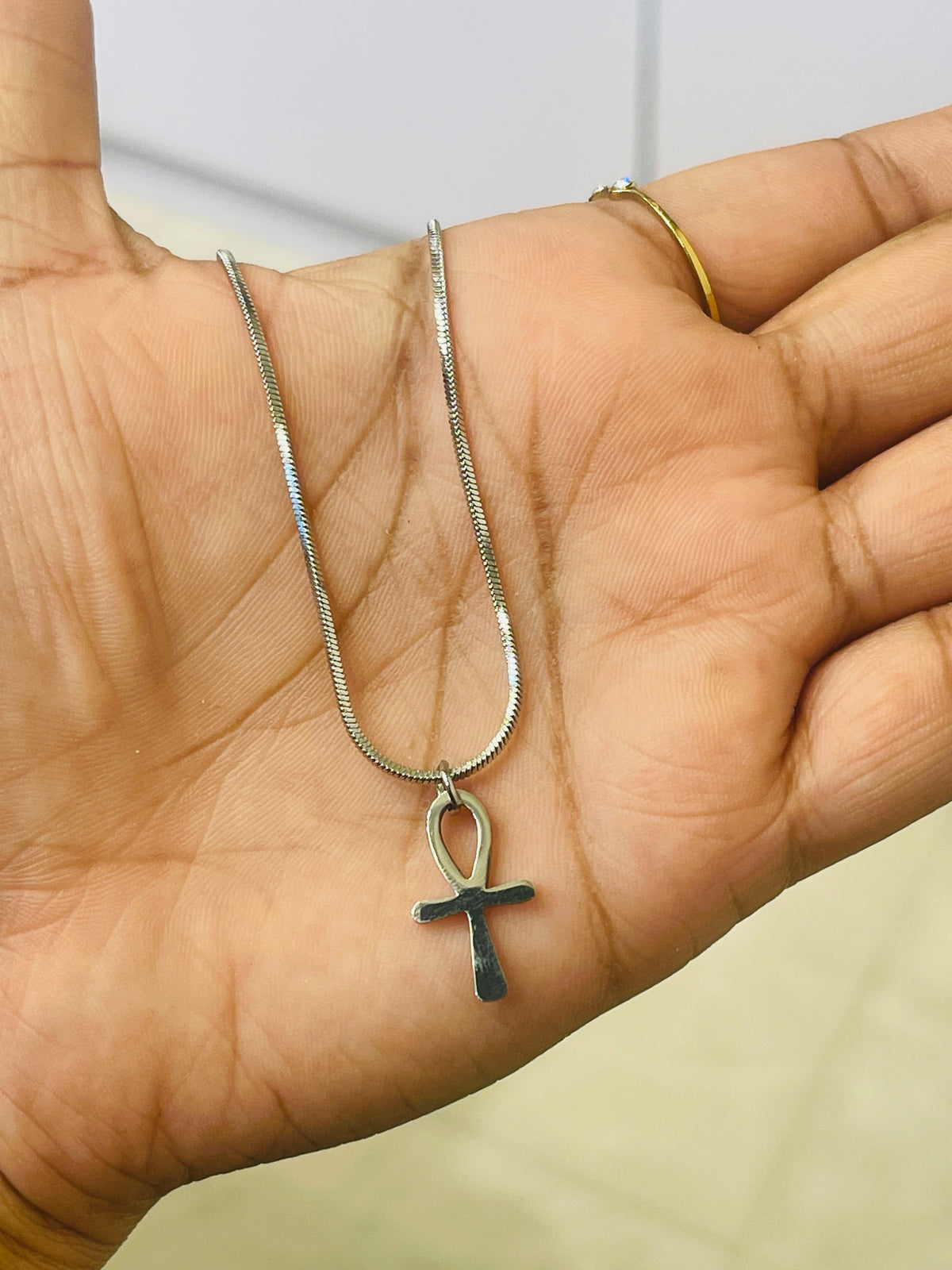 A silver snake chain necklace with a dark metallic ankh pendant, held in the palm of a hand. The person also wears a thin gold ring on their finger.