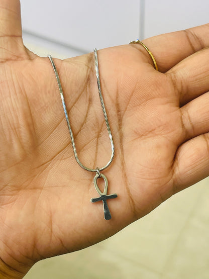 A silver snake chain necklace with a dark metallic ankh pendant, held in the palm of a hand. The person also wears a thin gold ring on their finger.