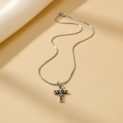 A silver cross pendant necklace with a detailed crucifix design, resting on a warm beige surface with soft shadows.