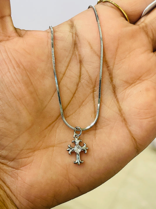 A silver cross pendant necklace with a clear central gemstone, resting on an open hand. The necklace is on a silver snake chain.