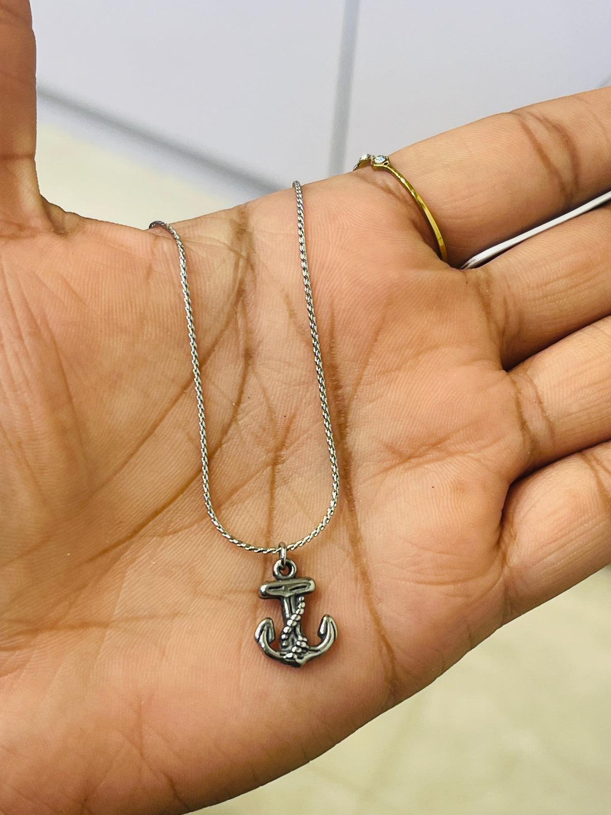 A silver anchor pendant necklace on a matching chain, held in a person's open palm. The person is wearing a thin gold ring on their index finger.