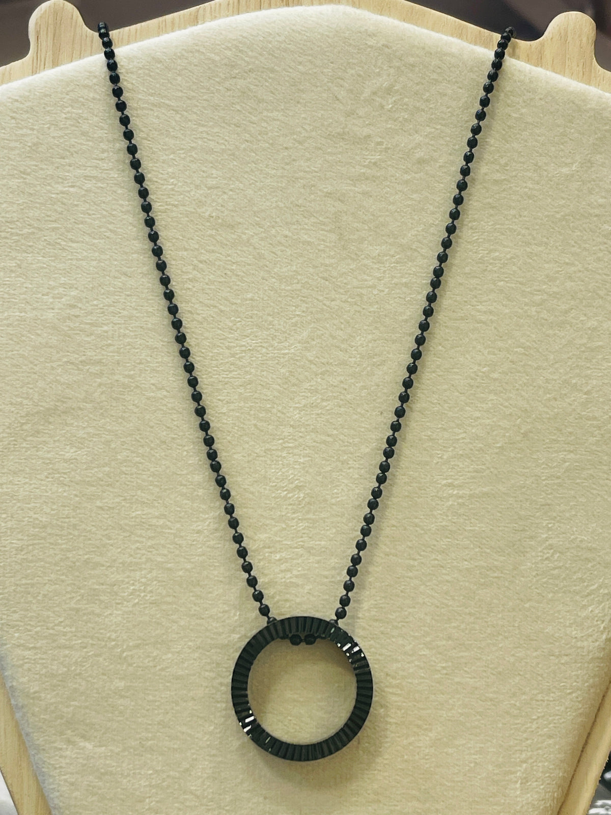A black beaded necklace with a textured circular pendant, displayed on a cream-colored jewelry bust.