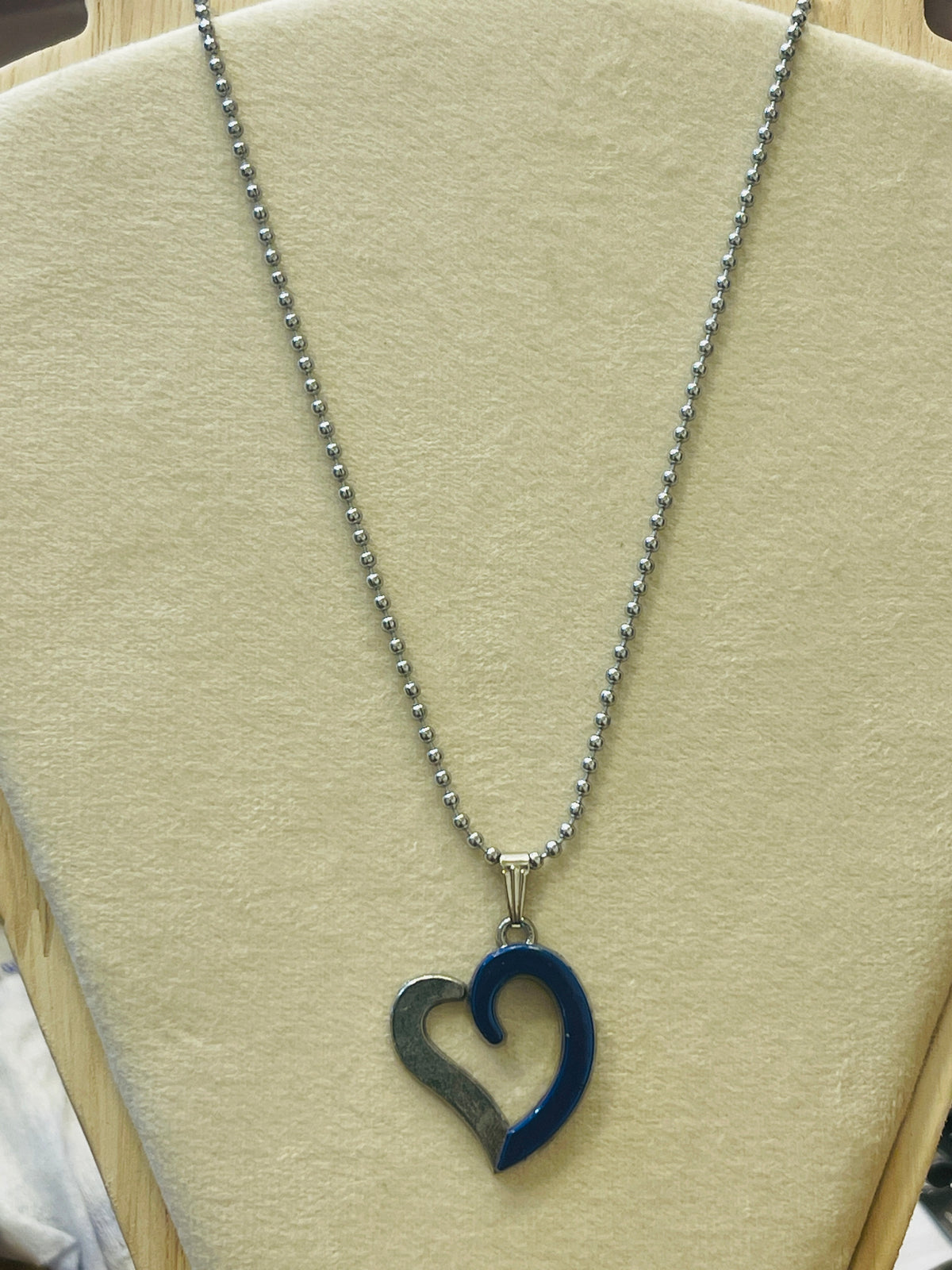 A silver ball chain necklace with a heart-shaped pendant, featuring one side in polished silver and the other in dark blue enamel, displayed on a beige jewelry bust.
