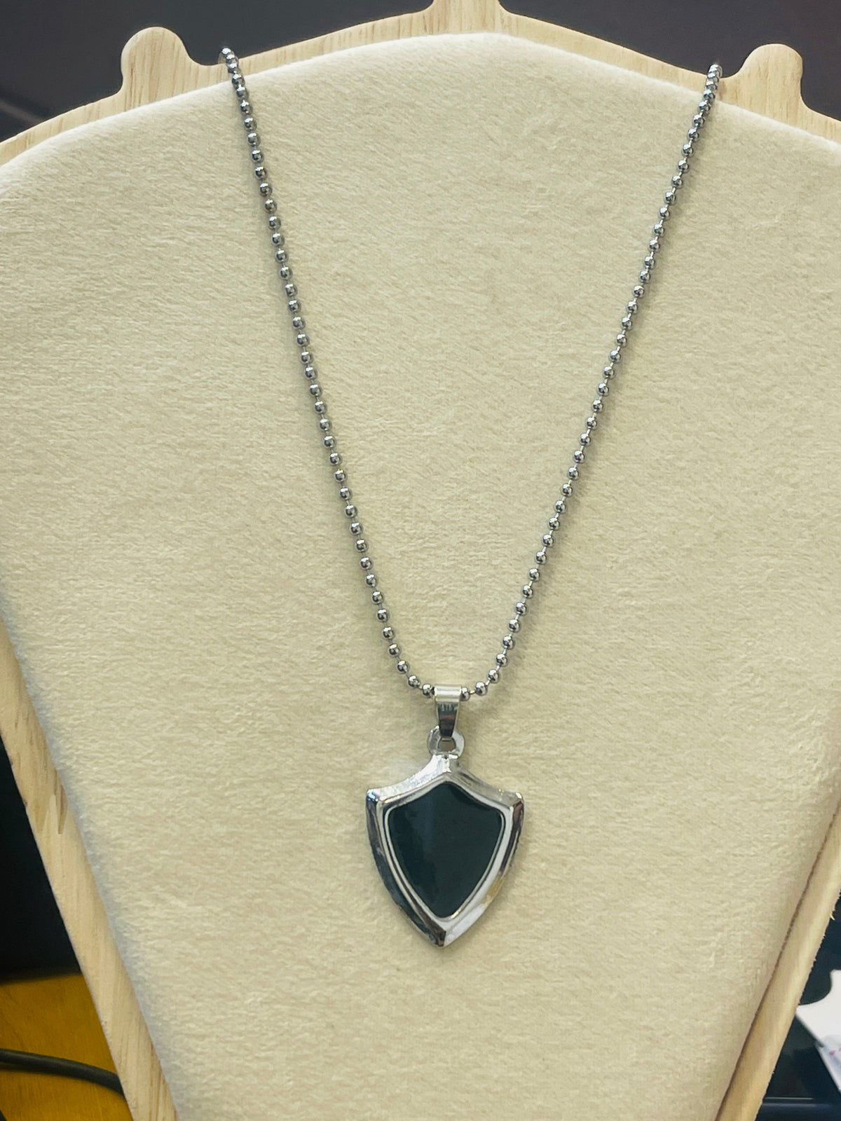 A silver-tone shield-shaped pendant necklace with a dark green inlay, displayed on a cream-colored jewelry bust.