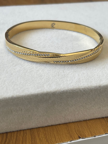 A gold bangle bracelet with a textured chevron pattern and a row of small clear stones, resting on a white jewelry box.