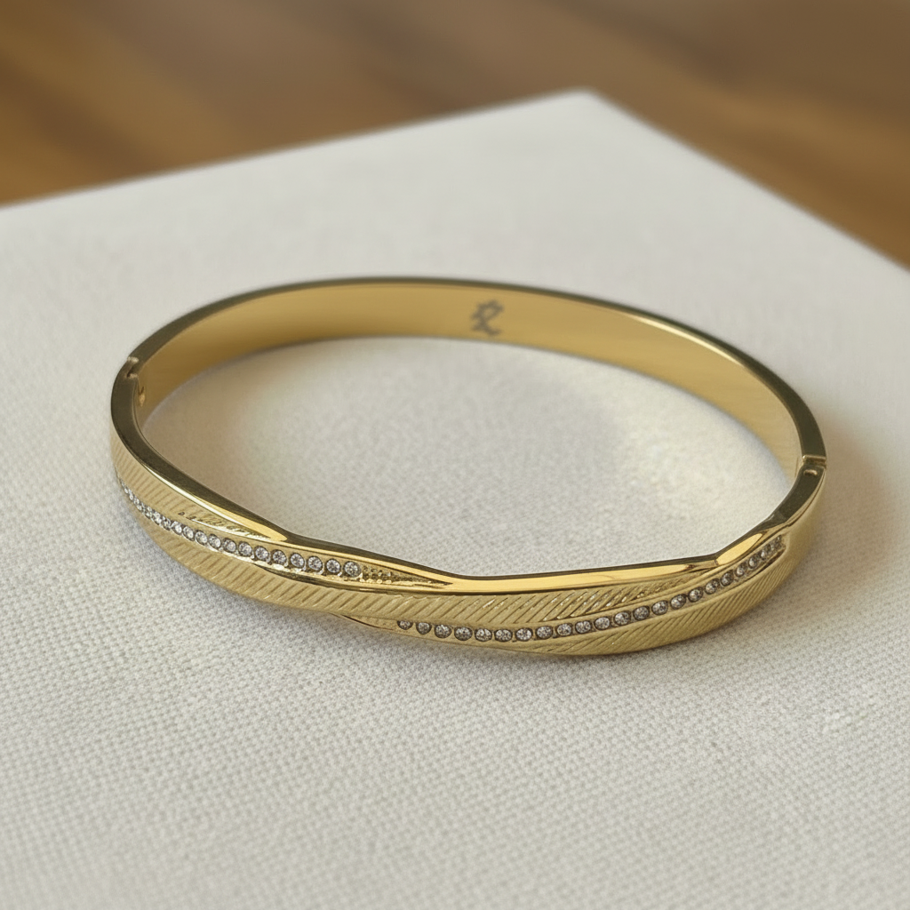 A gold bangle bracelet with a textured feather-like pattern and a row of small clear stones, resting on a white textured surface.