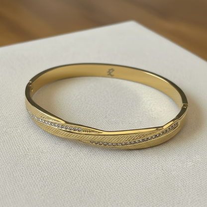 A gold bangle bracelet with a textured feather-like pattern and a row of small clear stones, resting on a white textured surface.