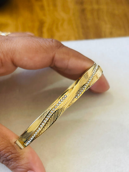 A gold-toned bangle bracelet with a textured chevron pattern and a row of small clear stones, held between fingers against a white surface with a blurred wooden background.