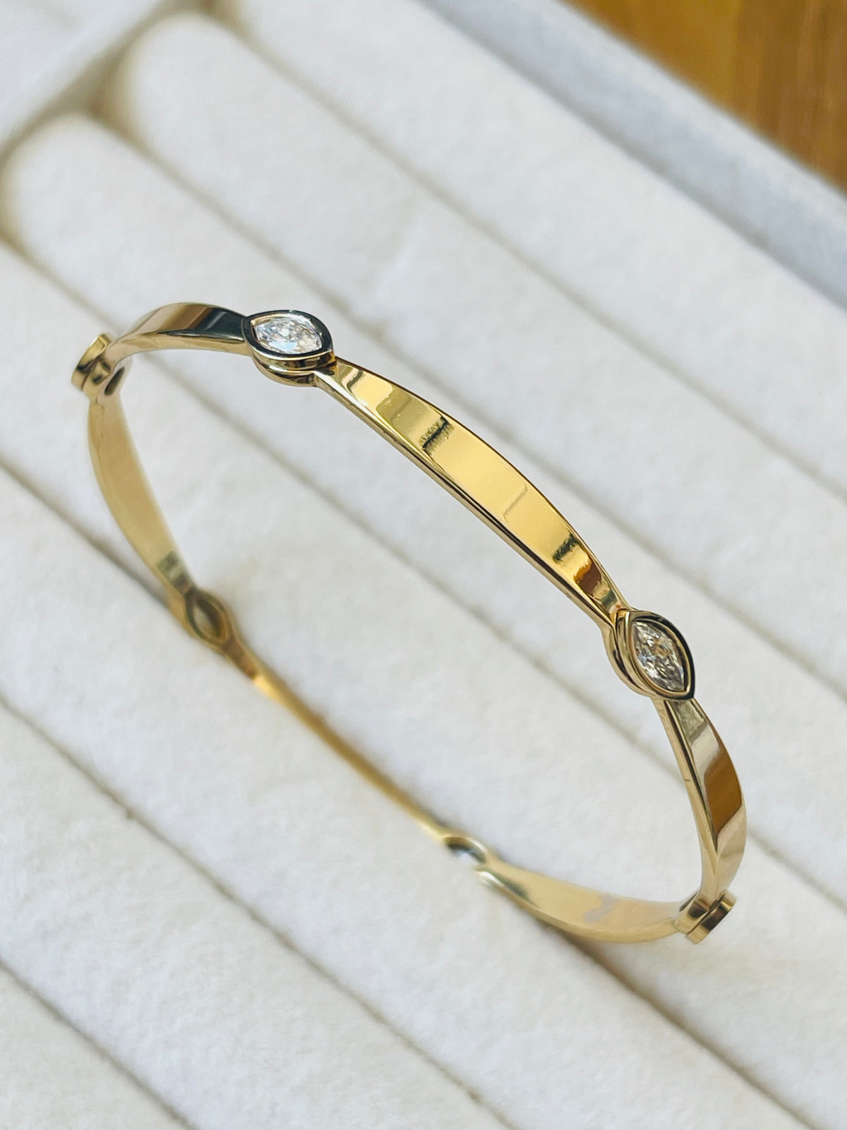 A polished yellow gold hinged bangle bracelet with two small marquise-cut clear gemstones, displayed on a white jewelry cushion.