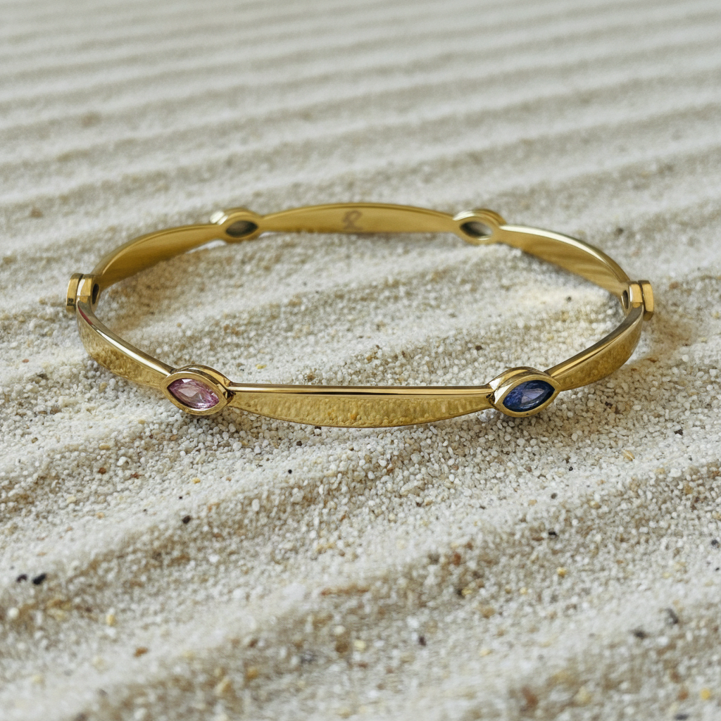 A gold-toned bangle bracelet with three small oval gemstone accents in pink, blue, and green, displayed on a bed of light beige sand.