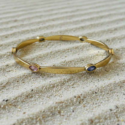 A gold-toned bangle bracelet with three small oval gemstone accents in pink, blue, and green, displayed on a bed of light beige sand.