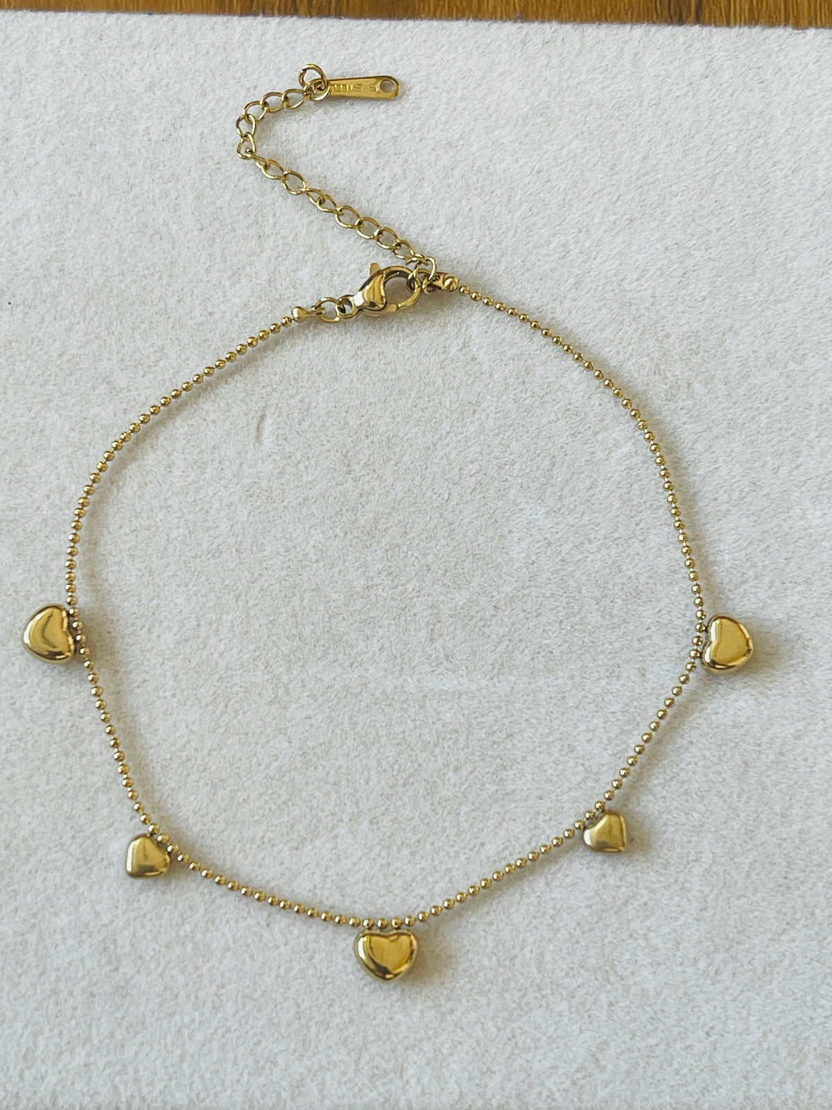 A gold chain anklet with five small heart charms, displayed on a textured white fabric background.