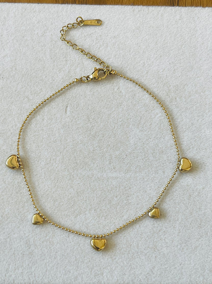 A gold chain anklet with five small heart charms, displayed on a textured white fabric background.