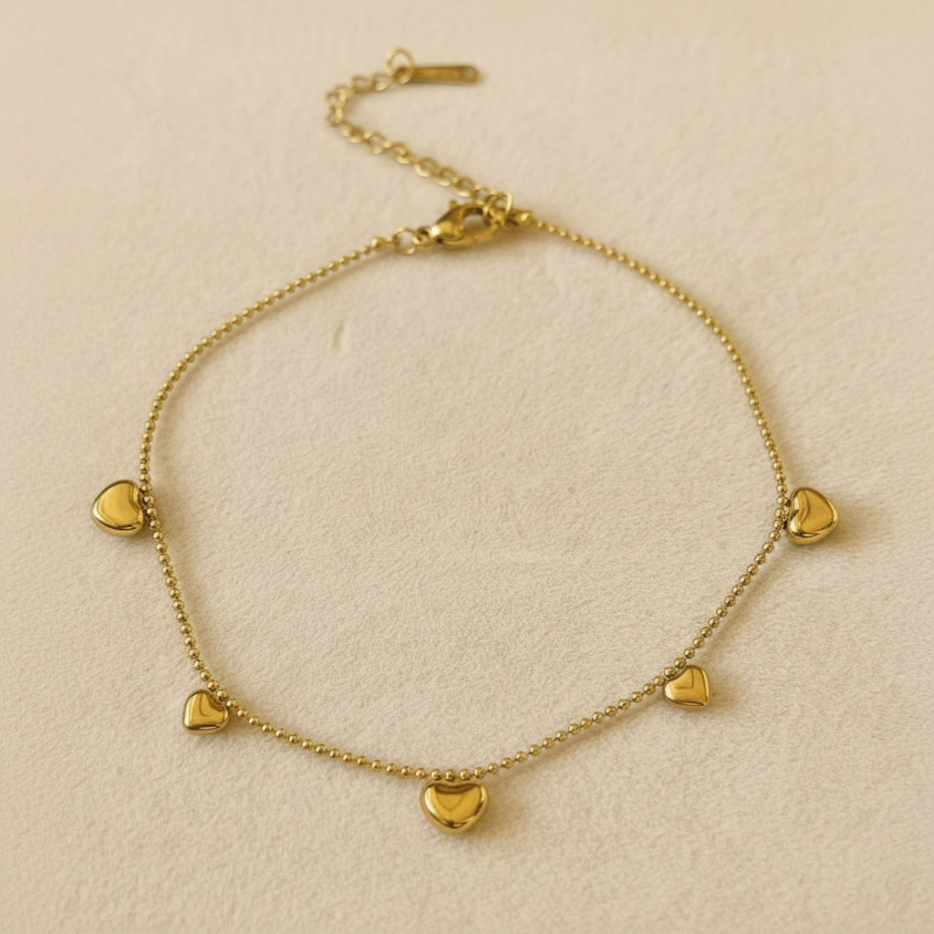 A delicate gold chain anklet featuring five small, polished heart charms, displayed on a textured beige fabric background.