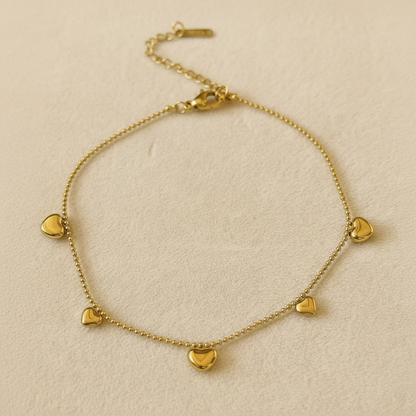 A delicate gold chain anklet featuring five small, polished heart charms, displayed on a textured beige fabric background.